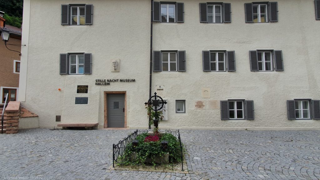 House in the background with the writing "Silent night museum Hallein", a grave in the foreground with an iron cross and greenary.