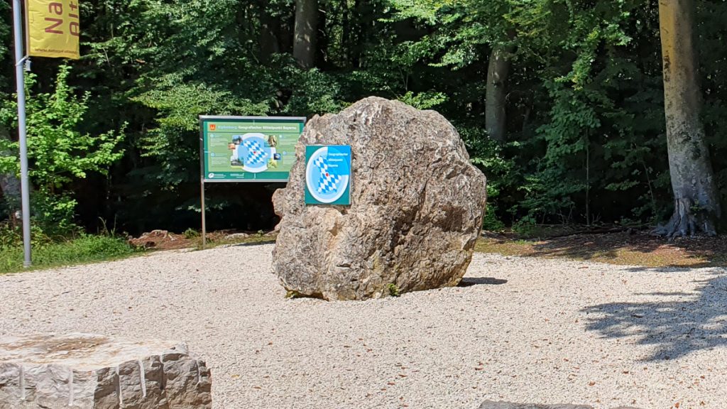 Crushed rock place surrounded by trees with a large rock in the middle having an information board attached to it
