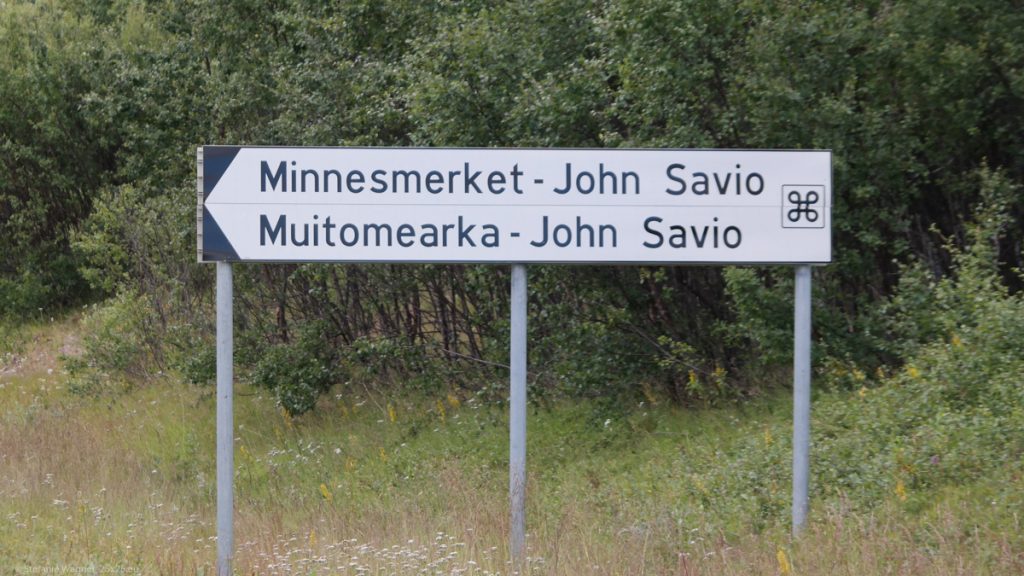 White street sign saying "Minnesmerket - John Savio" and the curved symbol next to it