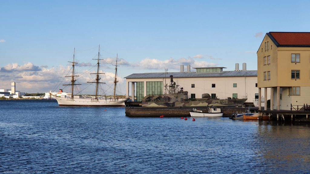 Long stretched building on the shore. A big white sailing board in the background, a modern camouflage military ship in the foreground.