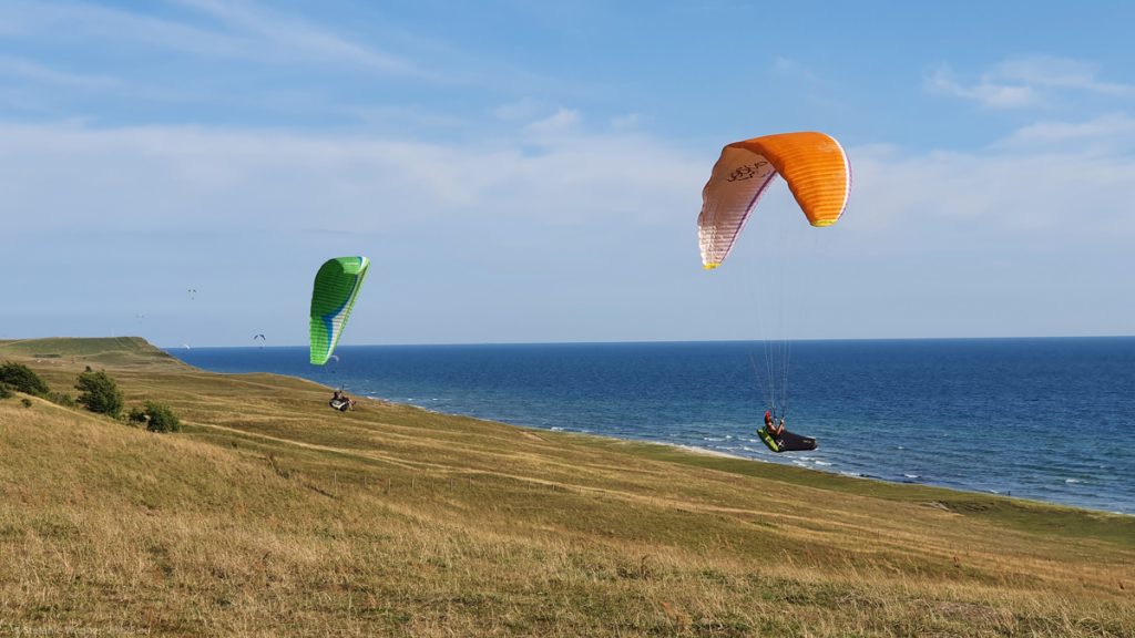 Two paragliders, one in the back with green parachute, one in the front with orange parachute. Being some meters above the ground. Ocean on the right side.