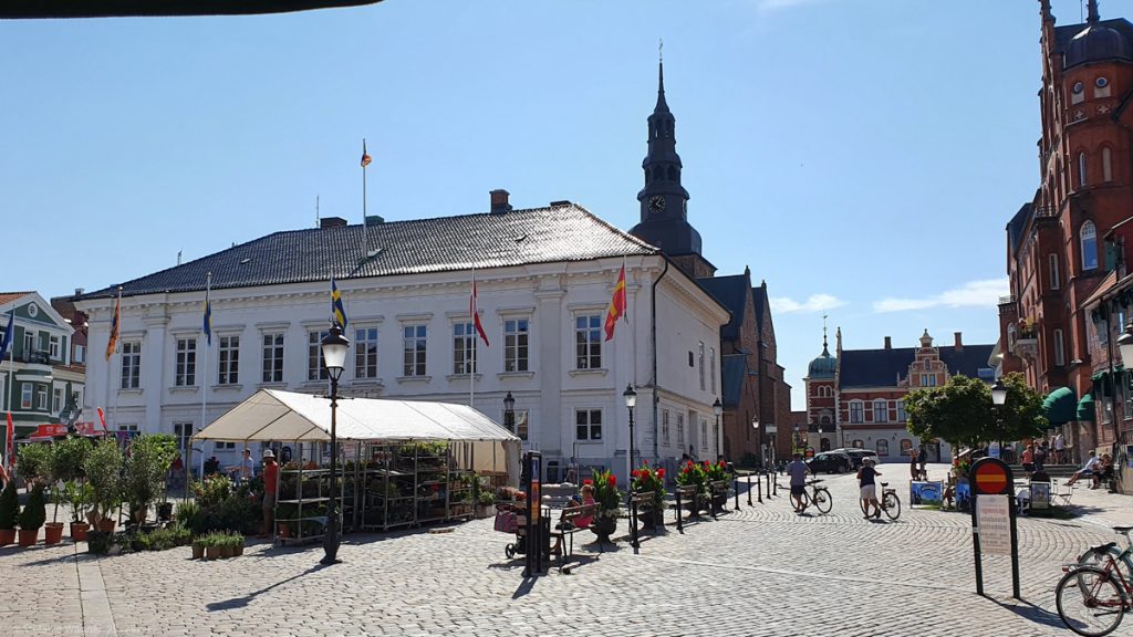 White building with ornaments, church tower in the background, red bigger building on the right, cobble stone place