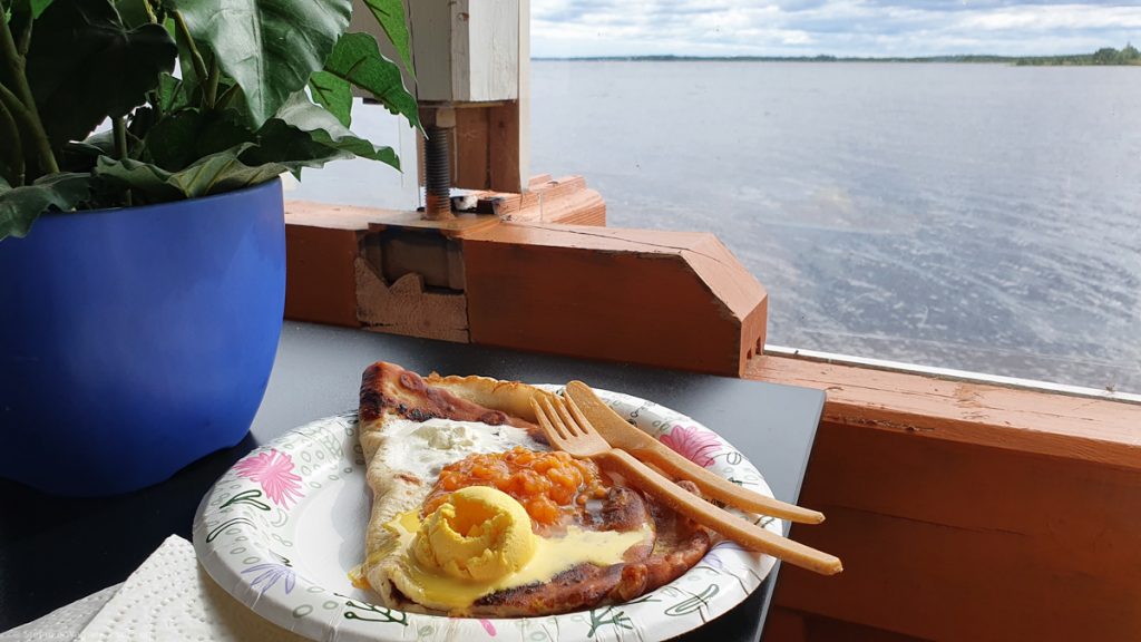 Paper plate with a folded pancake, on top some orange jam, yellow scoop of vanilla ice cream and some whipped cream, in the background a window with a lake