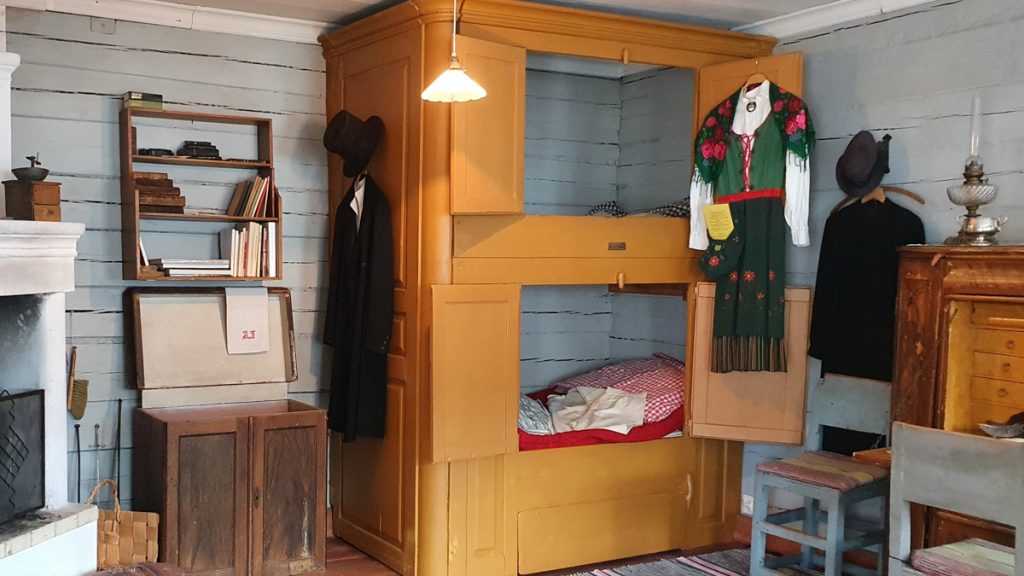 Inside a wooden house, brown cupboard with two levels, each of them with small doors. Inside one can see beds.