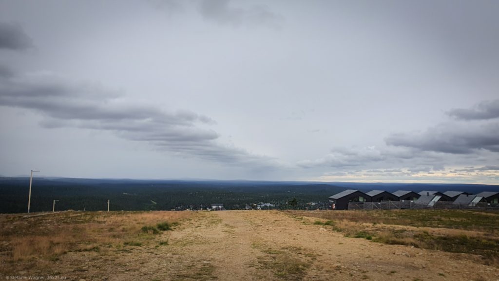 In the front brown soil, in the distance forests, on the right some small houses, almost all sky is cloudy besides some light on the right