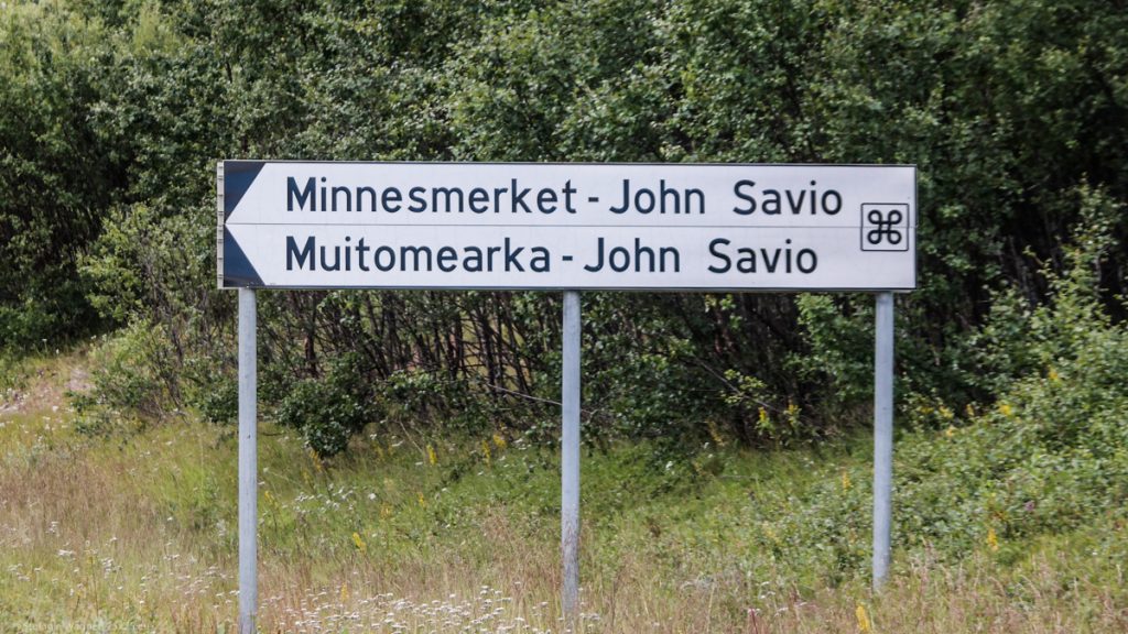 White sign saying "Minnesmerket - John Savio" at the side of the road