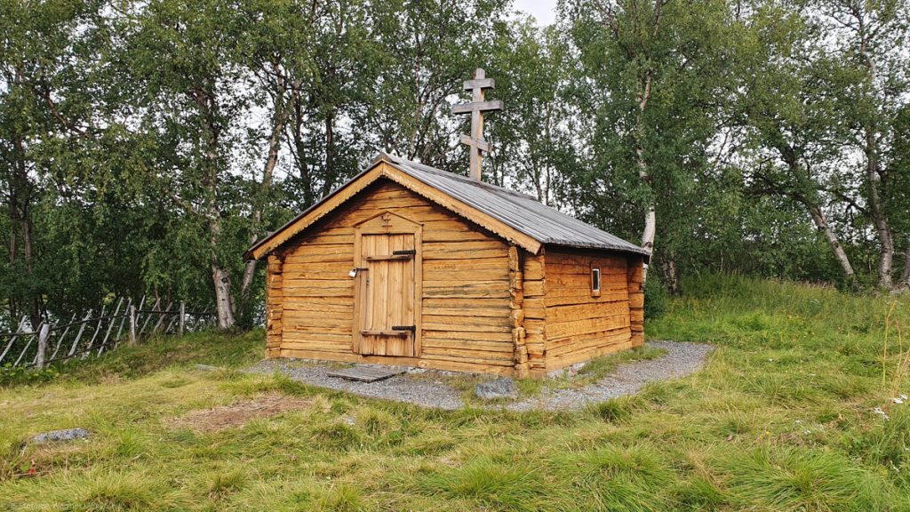 Small wooden building with light brown bars, one closed door at the front, one very small window on the side. Orthodox cross on the roof.