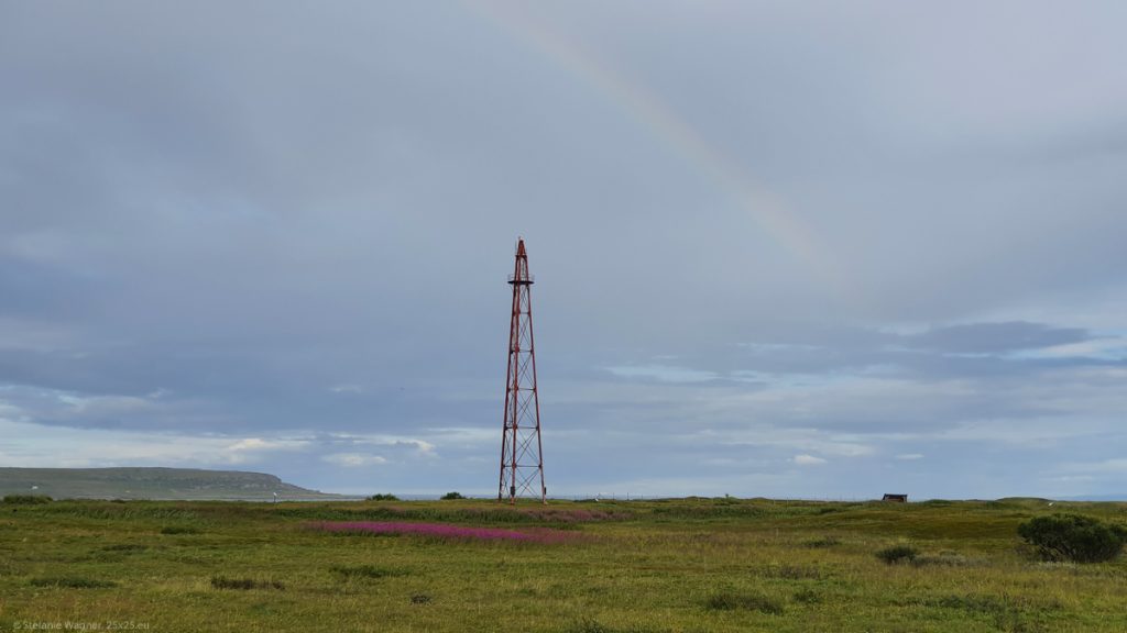 Red airship mast on a green meadow with some red flowers, dark clouds, part of a light rainbow visible