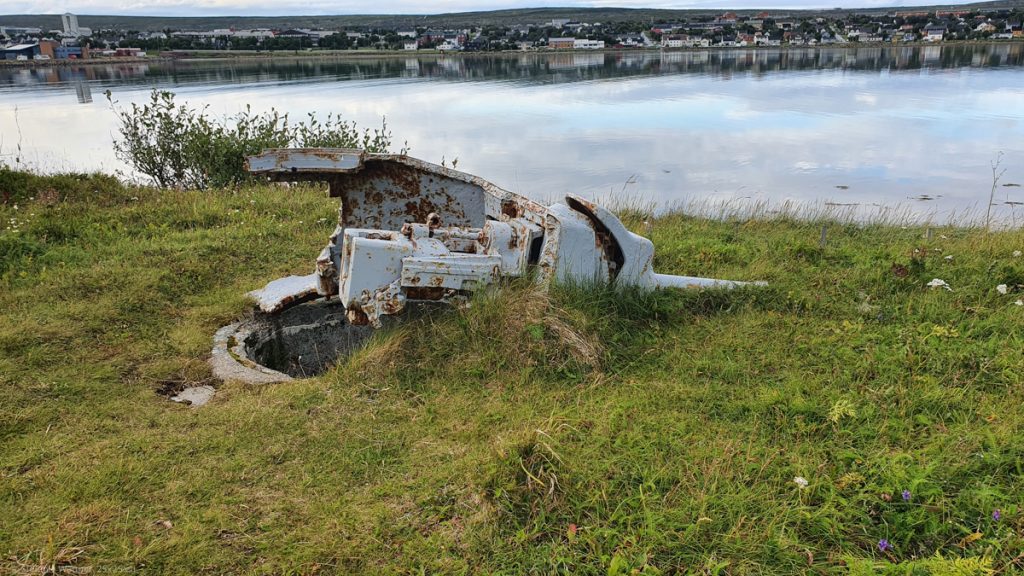 Remains of a gun emplacement. Gray, rusty metal structure lying on the grass, a concrete hole below, in the background water and the town