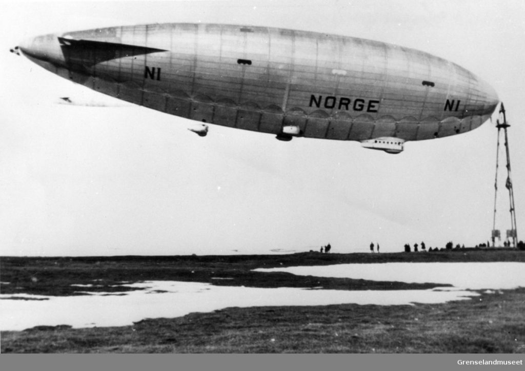 Black and white photo, air ship Norge in the air, tied to the air ship mast on the right, air ship mast is roughly a third of the length of the air ship