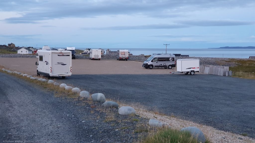 Gravel street on the left, parking lot style place with gray and reddish gravel on the right, some camper vans placed on it, in the far background the ocean